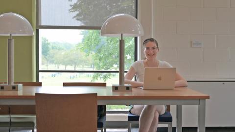 Elise Miller, a white, brown-haired woman with glasses and a ponytail sits behind a desk with a laptop on it.