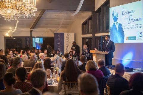 A photo of the Inaugural Keeper of the Dream dinner. Various tables with people sitting at them face a man speaking at a podium at the front of the room, and he stands in front of a projector screen depicting Martin Luther King Jr.