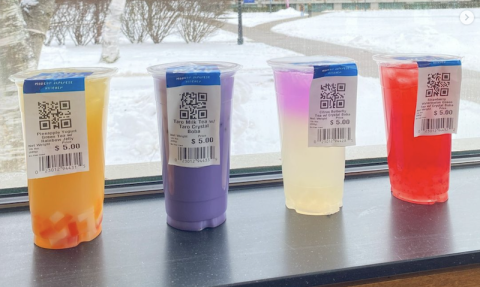 Four boba tea, orange, purple, clear, and red colored, rest on a black windowsill which overlooks the campus green. There's fresh snow on the ground and shrubbery in the background next to the stone walkway.