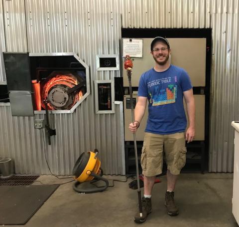 Brian Mernoff, an alum of the University. He is standing next to heavy machinery and is wearing a blue T-shirt and tan cargo shorts. He's smiling while holding a staff.