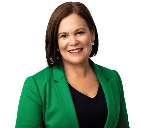 Mary Louise McDonald, president of Ireland’s Sinn Fein political party. She is in a green, long-sleeved jacket with a black undershirt. Her hair is brown and cut to chin-length, and she smiles at the camera against a white backdrop.