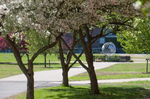 A stock photo of the campus globe. Several flowered threes border the walkway to the globe.