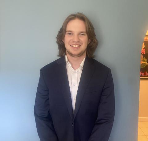 Austin Lourake, Class of 2025. He is in a blue coat jacket and white shirt and is posing for a headshot in front of a light blue wall.