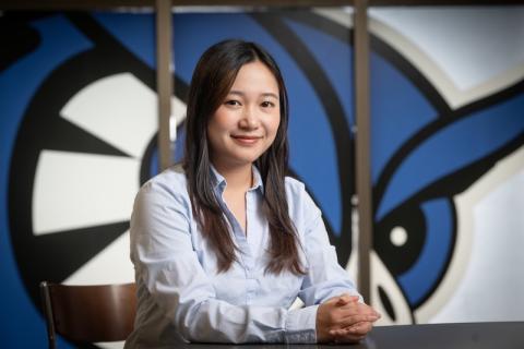 Honatsu Kabu, Class of 2025. A business administration major, Kabu wears a light blue shirt and sits at a desk in front of a Westfield State Owls mural.