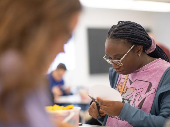 Student in pink shirt with glasses cutting paper in math classroom.