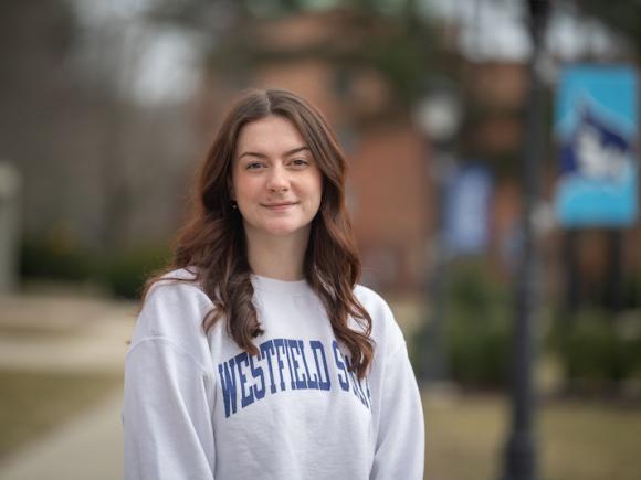 Paige Freeman, a senior at the University. She is outside, near the Ely building. She has long, brown hair and a gray sweatshirt which says "Westfield State" in blue lettering. Blurred out trees, grass, and the sidewalk are in the background.