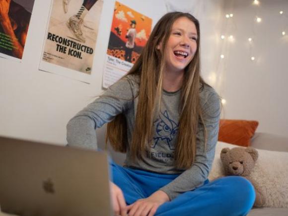 A student works on their laptop while sitting on their bed in their dorm room.