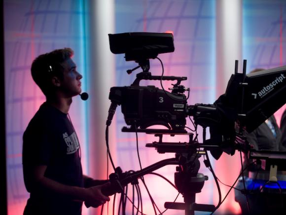 A student videographer during As School Match Wits. He is directing the competition through a stand-up camera and wears a mic around his ear and mouth.