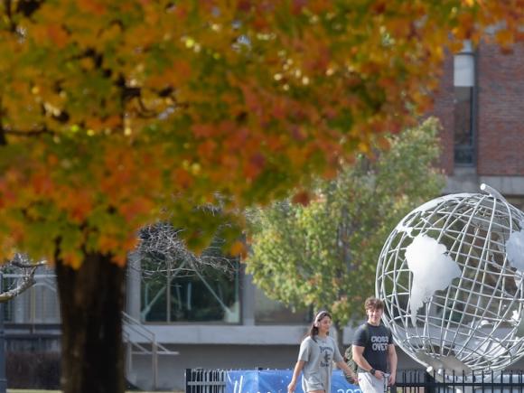 A stock photo of campus, with two students walking by the silver globe near Ely. In the corner of the photo is a tree with green and yellow leaves from autumn.