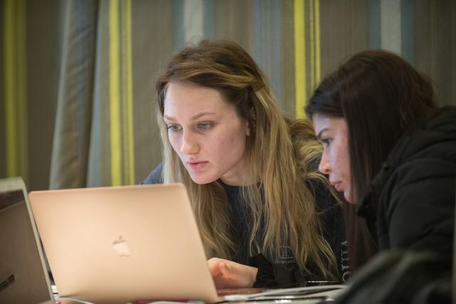 Student in front of laptop next to another student looking on. Yellow striped background behind them,