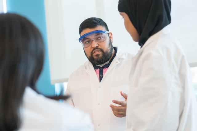 Chemistry faculty member wearing eye protection and white gown in a lab with two students.