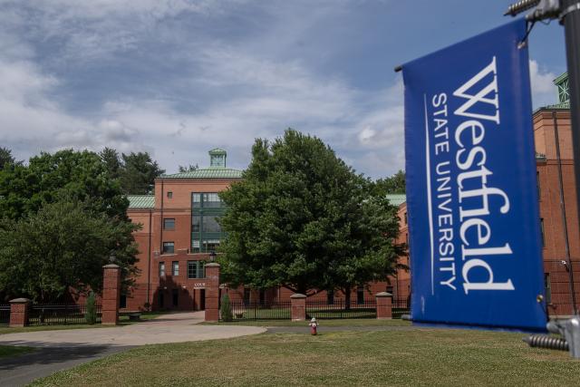 Courtney Hall with blue skies behind it and a Westfield State University blue and white flag in front of it.