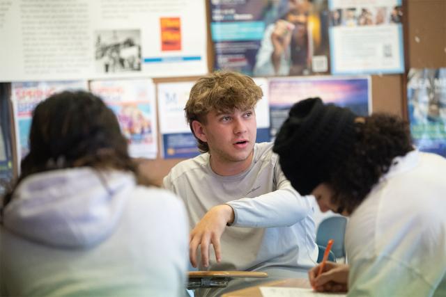 Three Westfield State University students sit in a group of desks while discussing and working on classwork.