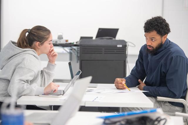 Two economics students sit at a desk discussing an assignment.