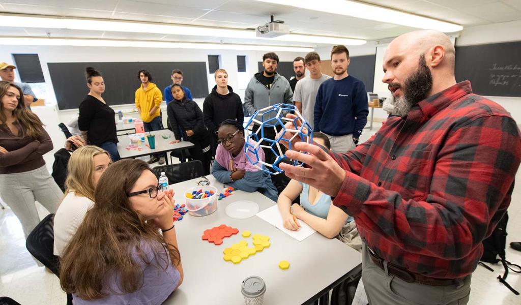 A math professor uses a 3D model to demonstrate mathematical concepts to students in a classroom.