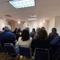 A shot of a student reading poetry at the Chapbook Presentation event on December 13. Rows of people sit in front in blue chairs, with the speaker reciting the poetry into a microphone.