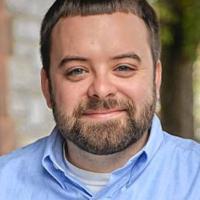 A close-up shot of Joe Courchesne, alumni of Westfield State and teacher at Granby Jr. and Sr. High School. He wears a light blue button-down shirt, and smiles at the camera with blurred out buildings and trees in the background.