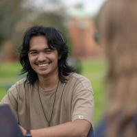 Riley Cosby '26, a communication major. He sits at a table on the campus green in a tan shirt and is laughing at something a friend is saying.